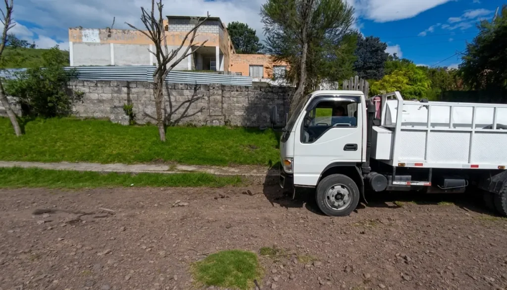 Imagen horizontal con efecto espejo de un camión cisterna de agua blanco sin marcas, estacionado en un camino de tierra. El camión está a la derecha, y el paisaje con una casa de dos pisos y árboles está a la izquierda, bajo un cielo azul con nubes.