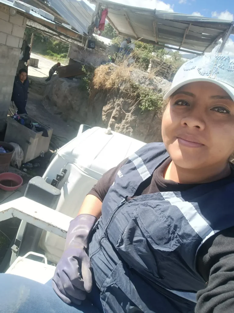 Selfie de una operaria del personal de limpieza vistiendo chaleco azul de trabajo con cintas reflectivas, guante de protección de goma y gorra clara, sentada en la parte posterior de un vehículo de servicio.