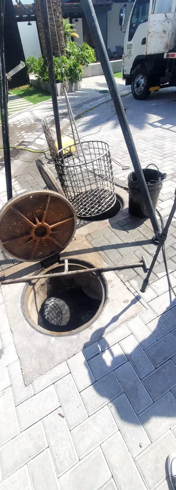 Vista en ángulo de una alcantarilla abierta en una acera pavimentada, con su tapa de hierro fundido oxidado a un lado y un trípode de elevación metálico encima. Una jaula cilíndrica de malla metálica está suspendida sobre el pozo por un cable, y un cubo de plástico negro lleno de material oscuro está cerca.