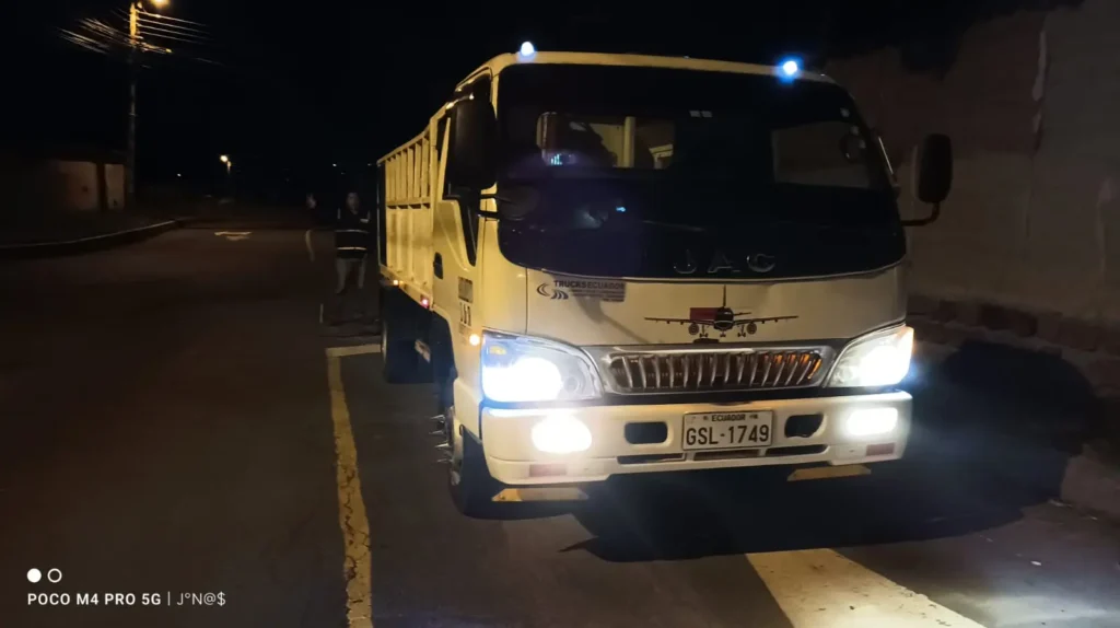 Nosotros contamos con un camión de plataforma blanco JAC con faros brillantes y luces superiores azules encendidas, estacionado de noche en una calle de Ecuador.