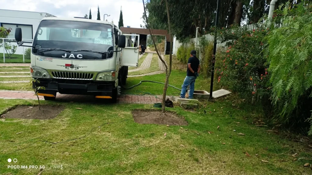 Camión cisterna de saneamiento realizando trabajos de succión y limpieza de pozo séptico en un jardín residencial.