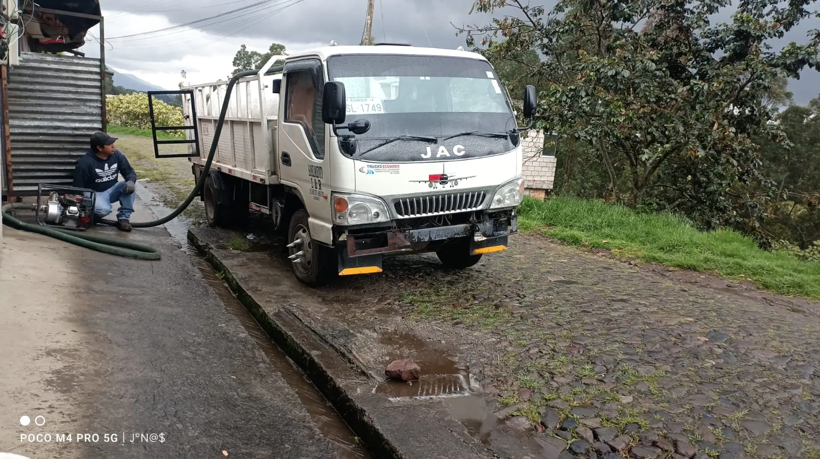 Camión cisterna blanco marca JAC estacionado en un camino de empedrado junto a un operario que opera una motobomba con mangueras de succión.
