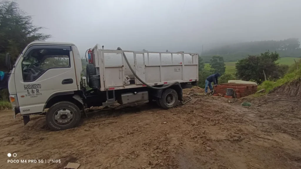 Camión cisterna de saneamiento realizando una atención de emergencia 24/7 en un terreno embarrado bajo un clima neblinoso, mientras un operario maneja la manguera de succión.