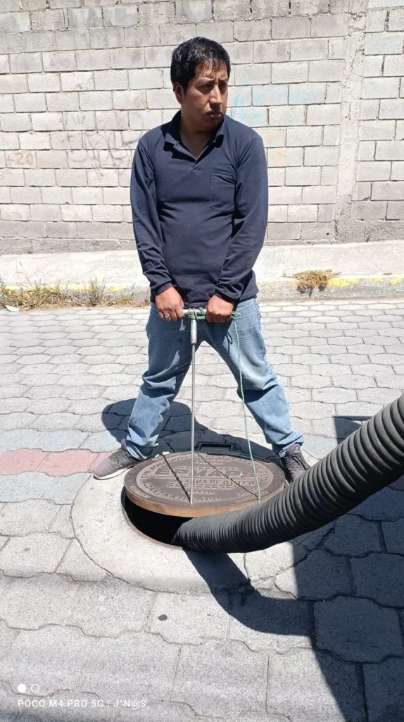 Operario utilizando una llave T de metal para levantar una tapa metálica de alcantarilla mientras una manguera de succión negra se introduce en el pozo.